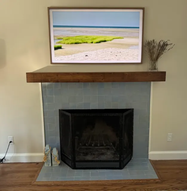 Living room fireplace with a framed landscape photograph of a beach scene, a wooden mantle, and a vase with dried plants on the mantle.