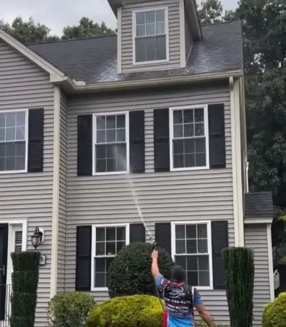 A person uses a pressure washer to clean the exterior of a two-story gray house with black shutters and landscaped bushes.