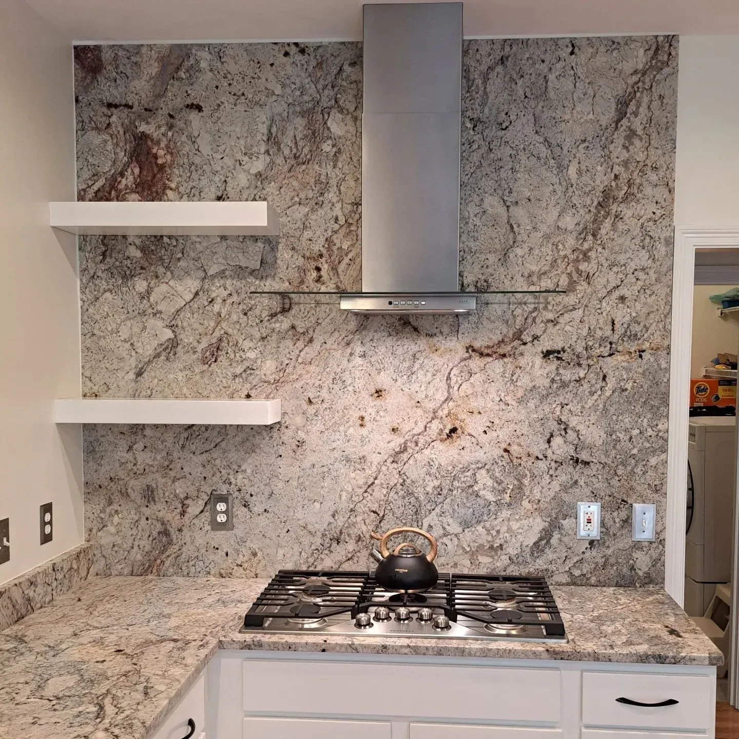 Kitchen backsplash with granite vs, a stainless steel range hood above the stove, white floating shelves to the left, and a black kettle on the stove.