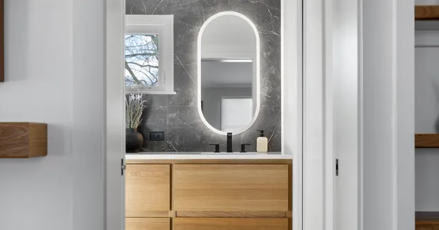 Modern bathroom vanity with natural wood cabinetry, illuminated oval mirror, black fixtures, and under-cabinet toe-kick lighting, viewed from a hallway entrance.