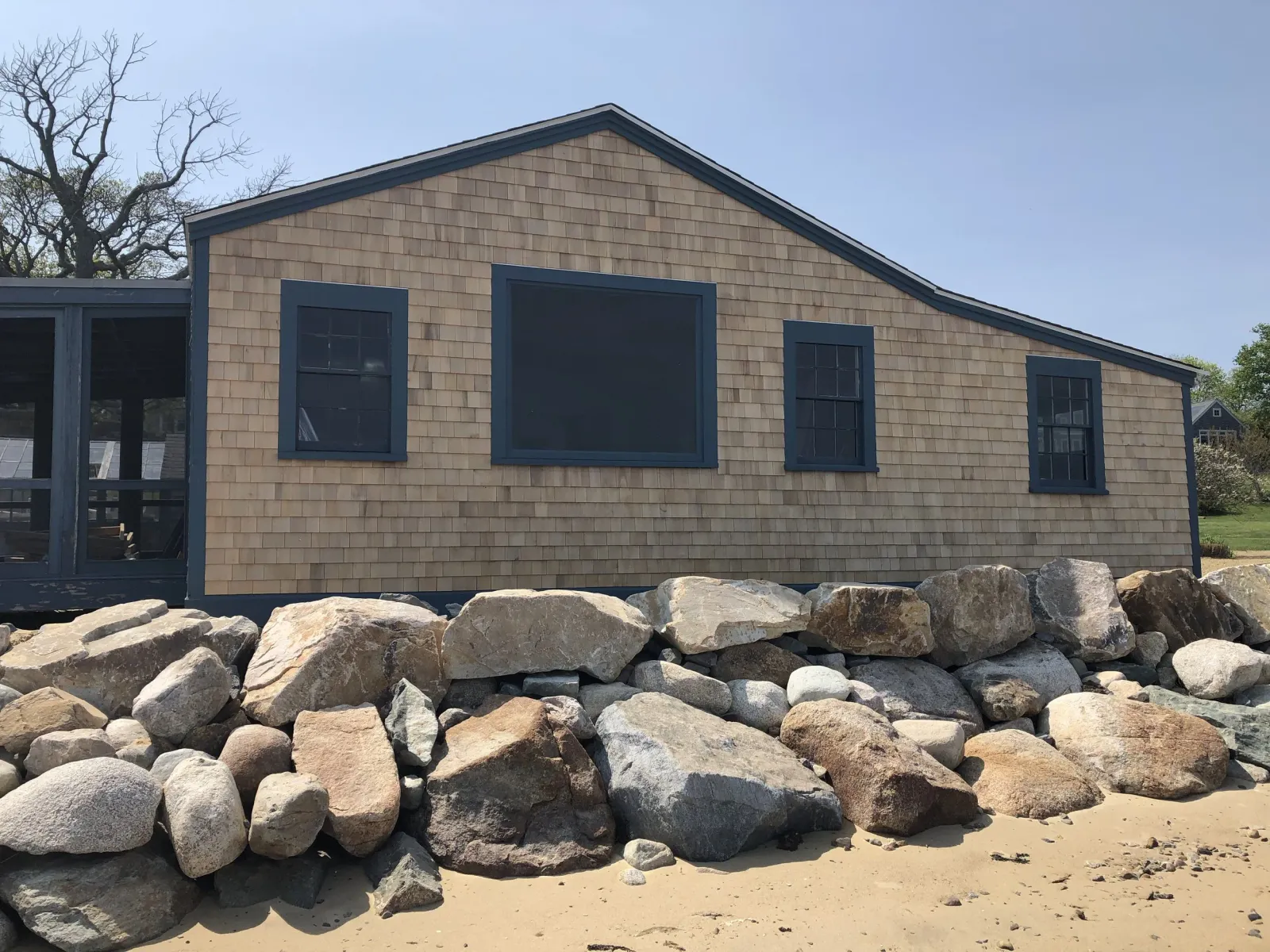 Beach house with shingle siding and dark blue trim, situated on sandy beach with large rocks in foreground, clear sky in background.