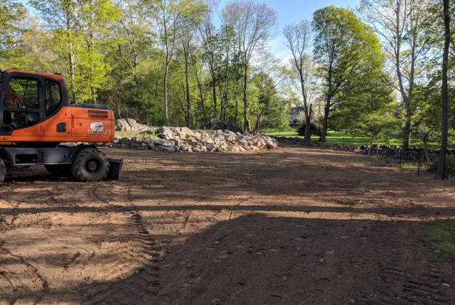 Orange excavator on a dirt clearing beside a wooded area under a bright sky