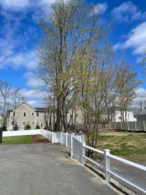 Driveway with white fence, trees, grass, and two-story houses under a partly cloudy blue sky.