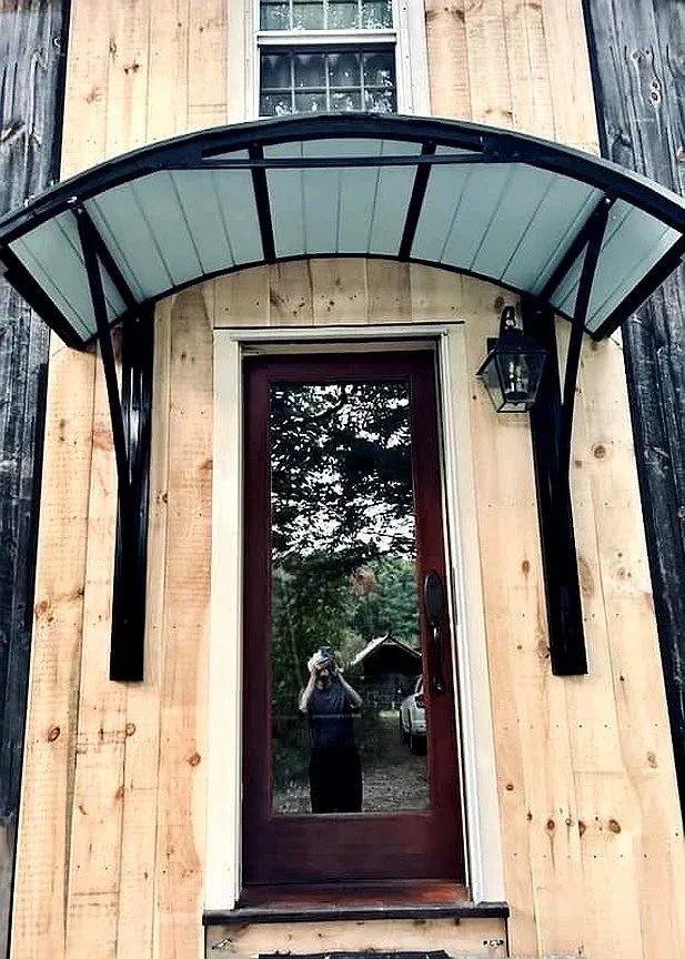 A front door with a glass storm door inside a wooden wall. A black metal or metal-like awning extends above the door, supported by two black brackets. A black outdoor wall lantern is mounted beside the door. A reflection of a person taking the photo