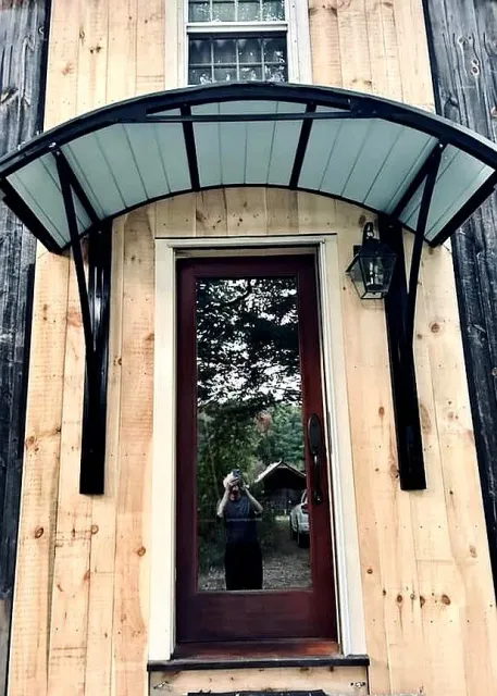 A front door with a glass storm door inside a wooden wall. A black metal or metal-like awning extends above the door, supported by two black brackets. A black outdoor wall lantern is mounted beside the door. A reflection of a person taking the photo