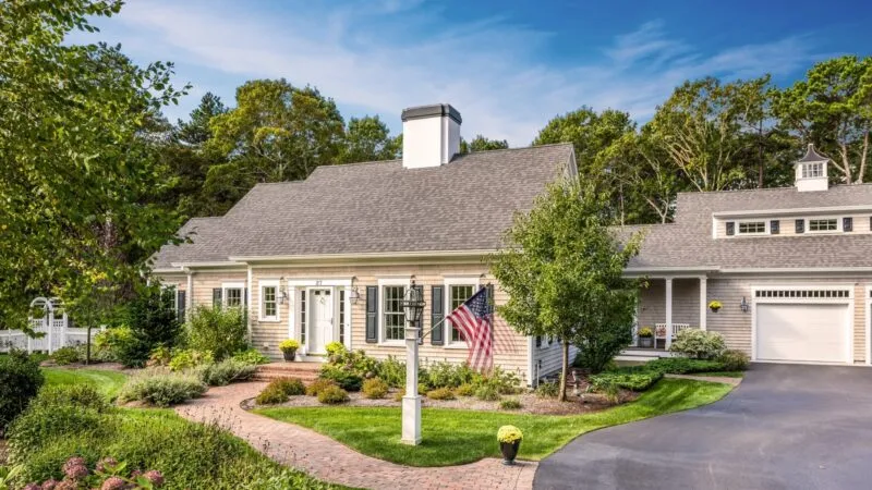 Charming single-story home with gray siding, white trim, brick walkway & flag – McPhee Associates of Cape Cod.