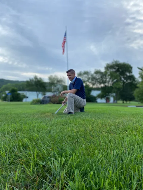 Man kneeling on grass field smiling with an American flag blurred in the background under a cloudy sky.