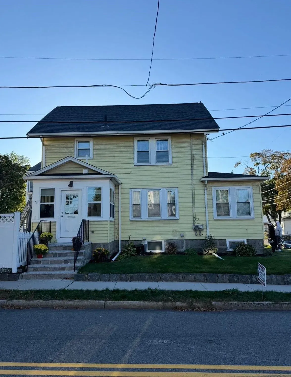 Yellow two-story house with a porch, white trim, and dark roof beside a street on a clear day
