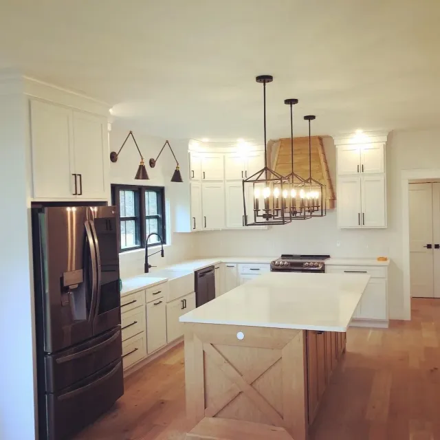Modern kitchen with white cabinets, black handles, a stainless steel refrigerator, a black dishwasher, a black stove, a kitchen island with a wooden base and white countertop, black pendant lighting, a window above the sink, and wood flooring.