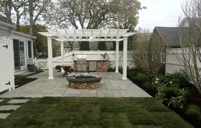 Backyard patio with white pergola, fire pit, stone paving, and landscaped garden beds.