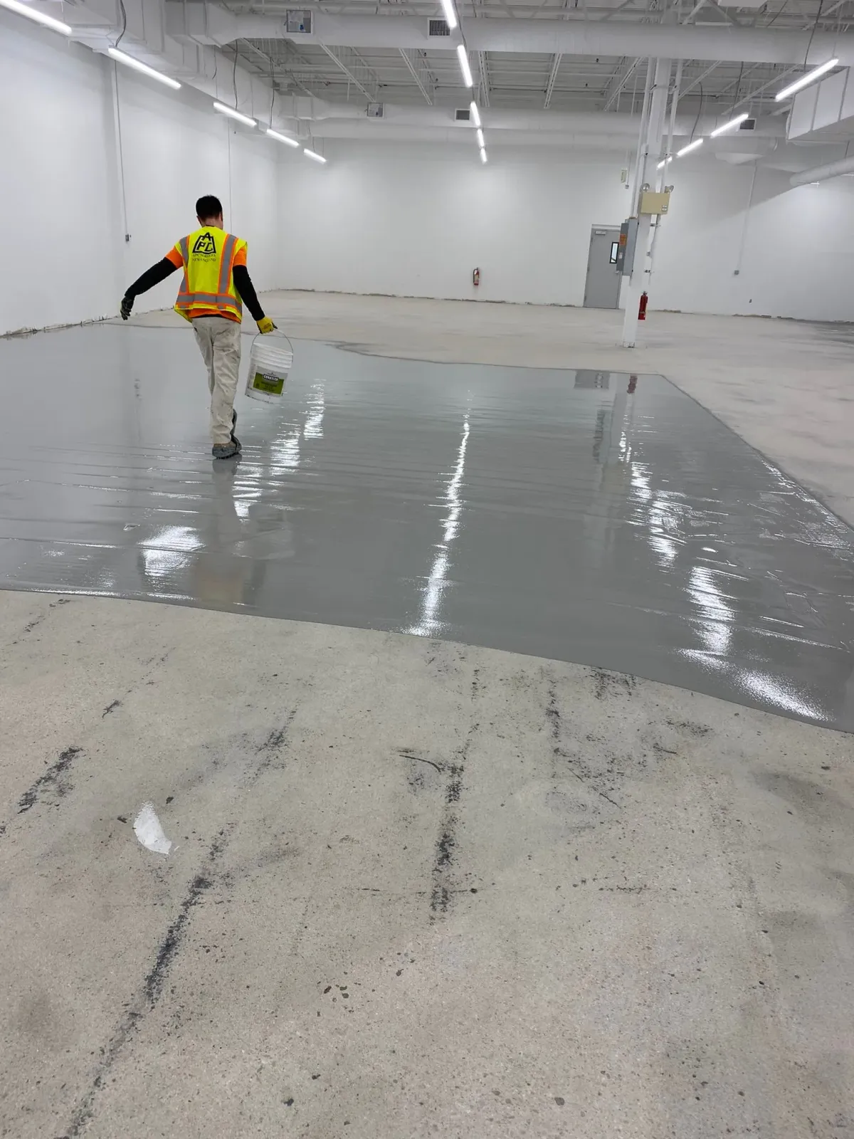 Worker pouring or smoothing gray floor coating in a large empty warehouse interior