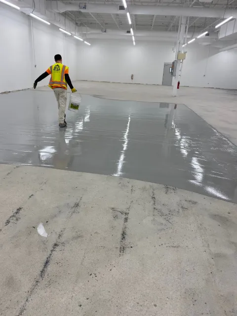 Worker pouring or smoothing gray floor coating in a large empty warehouse interior
