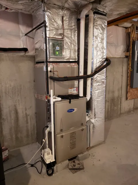 A heating, ventilation, and air conditioning (HVAC) system installed on a concrete basement floor in a building, with various pipes, electrical components, and insulation around it.