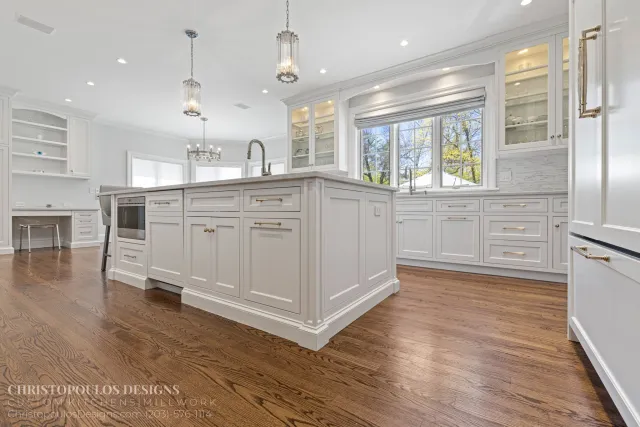 A bright, modern kitchen with white cabinets, a large island, hardwood floors, and three pendant lights hanging from the ceiling. Large windows offer a view of trees outside.