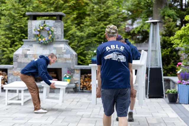 Group of people moving outdoor patio furniture with a stone fireplace, flowers, and trees in the background.