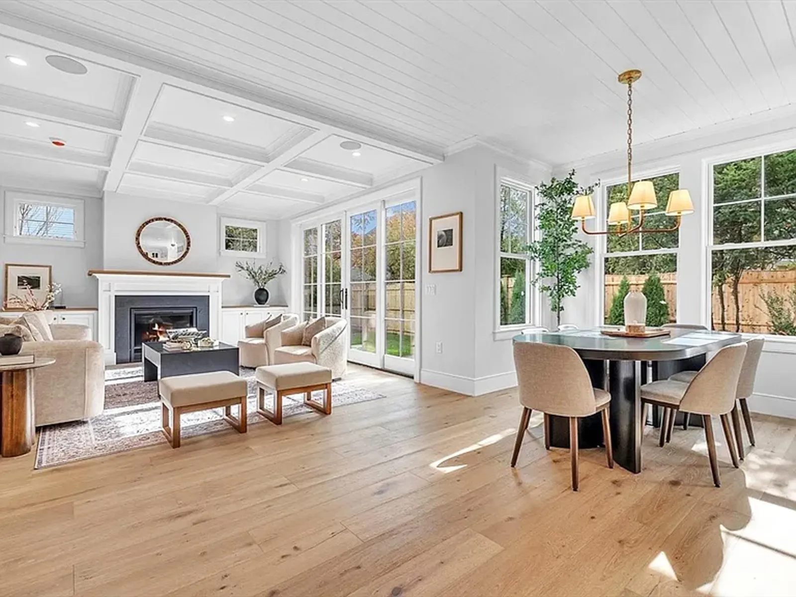 Hardwood floor installation in a Norwell living and dining room featuring white walls, large windows, a fireplace, a beige sofa with matching armchairs, a black coffee table, and a round dining table with six chairs under a chandelier.