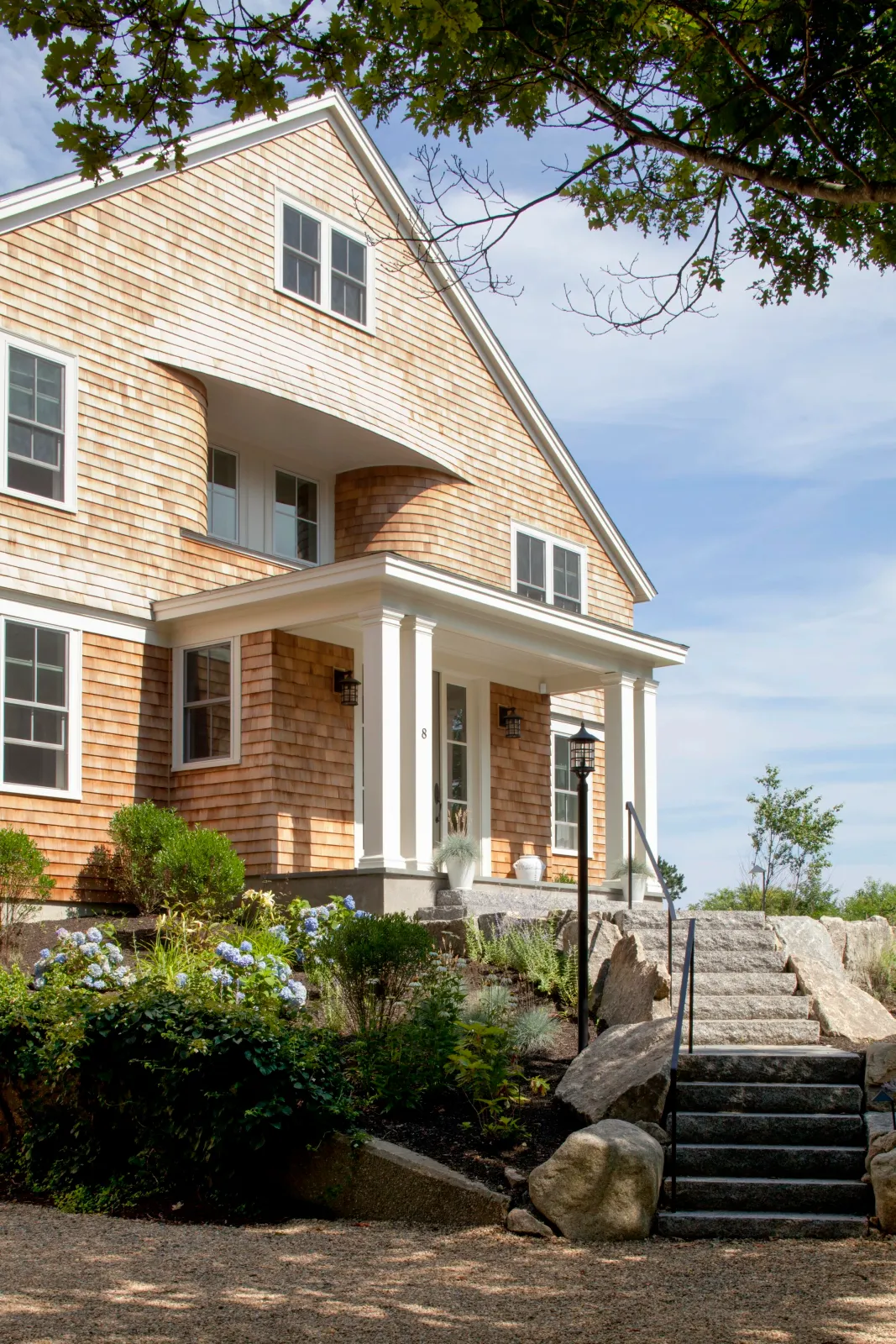Front exterior of a large, modern house with a porch, white columns, and stone steps leading up to the entrance, surrounded by landscaped plants and rocks under a blue sky.