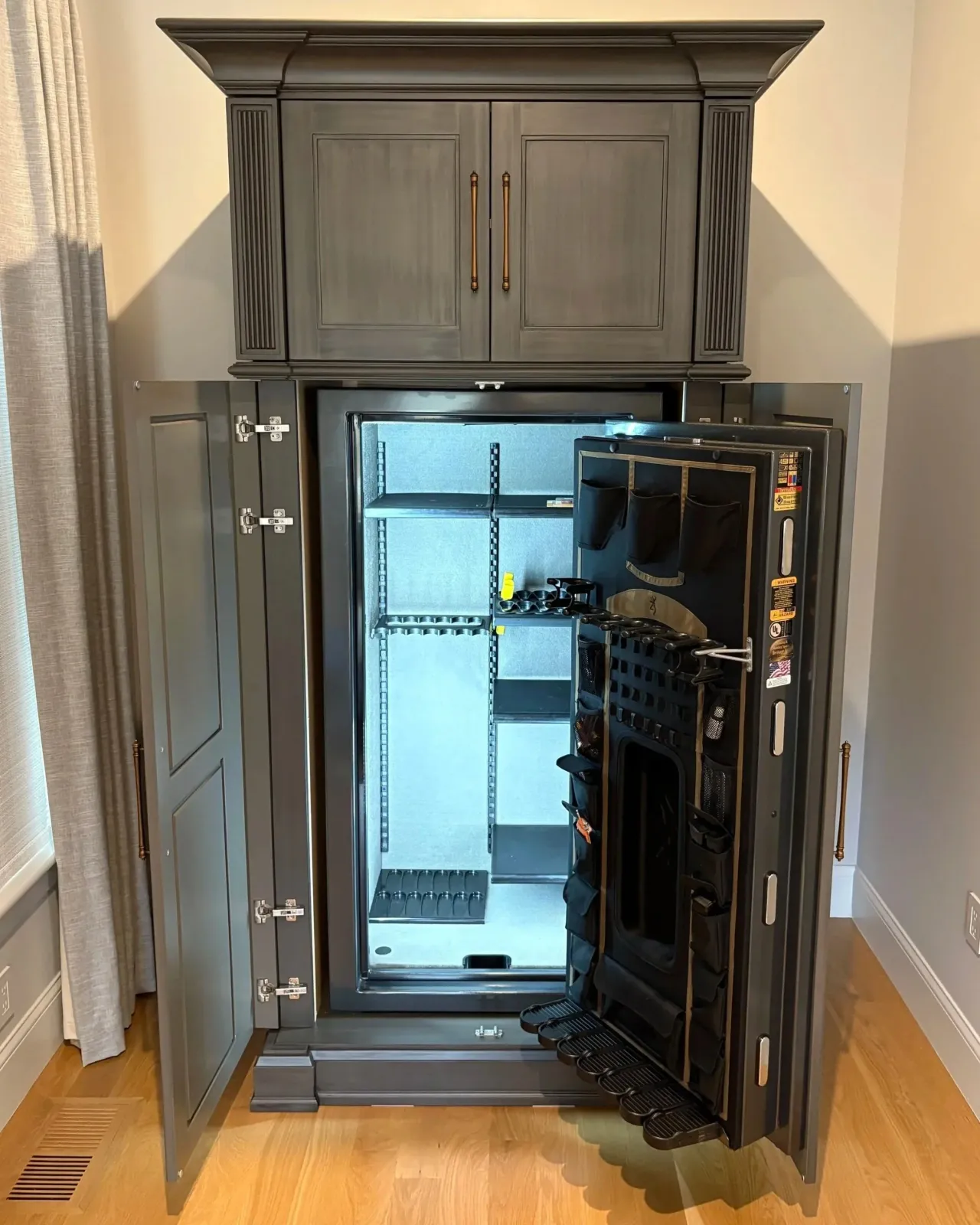 Open black safe with shelving and storage compartments, located in a room with wooden flooring, beige walls, and a baseboard heater. A black tool organizer is hung on the right side of the open safe door.