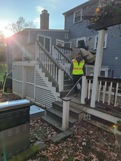 A man wearing a yellow safety vest standing on a wooden deck staircase of a gray house, with a large chimney, a clear sky, fallen leaves on the ground, and outdoor furniture piled on the deck.