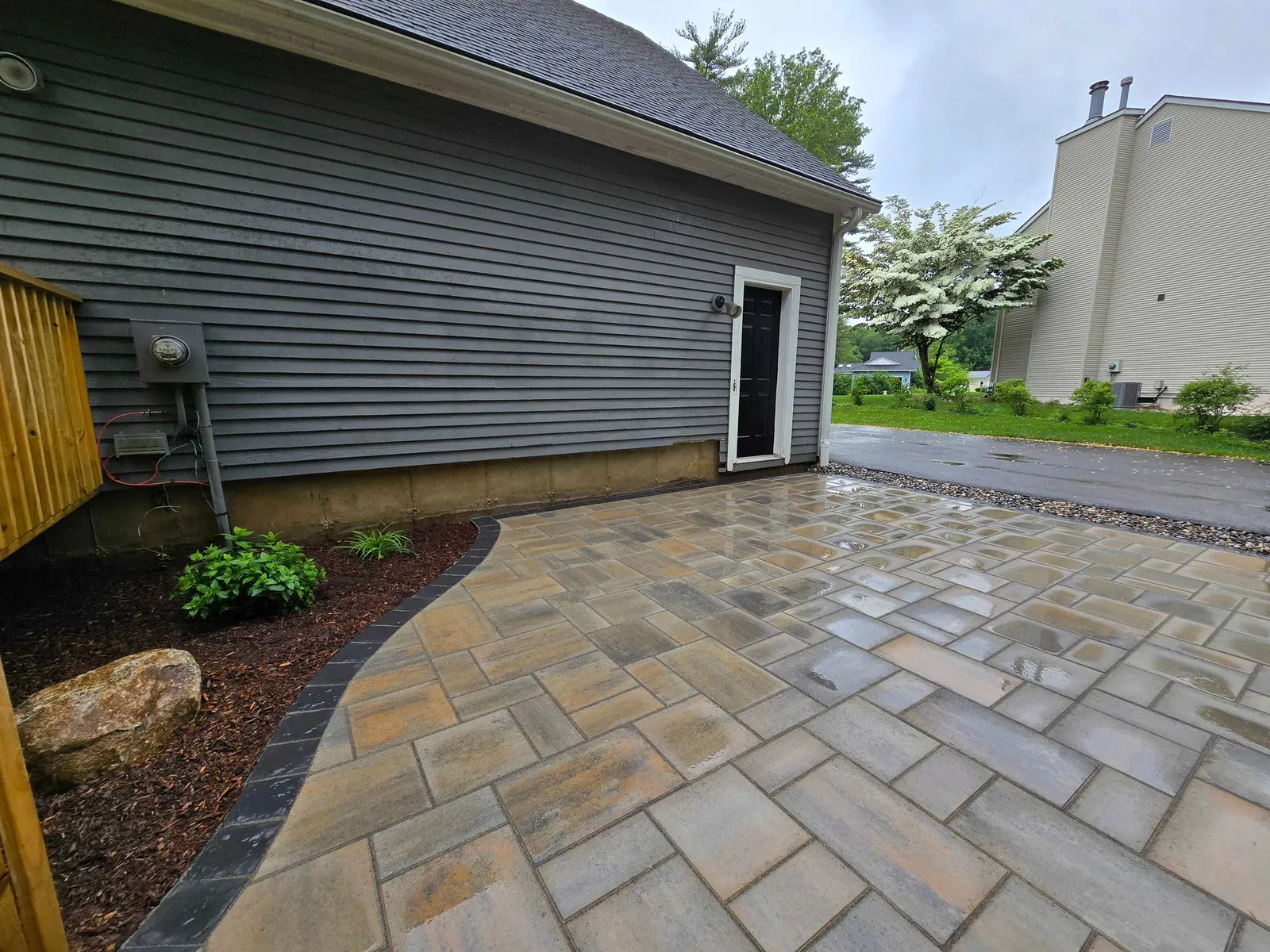 A gray-sided house with a newly installed stone patio and a dark stone border, next to a small garden bed and a driveway.
