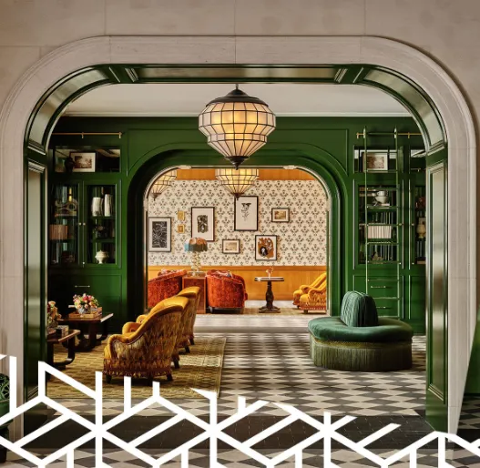 Interior of a vintage-style living room with green walls, ornate arched doorway, patterned wallpaper, and antique furniture including velvet armchairs, a round green sofa, and wooden tables, illuminated by a large pendant light.