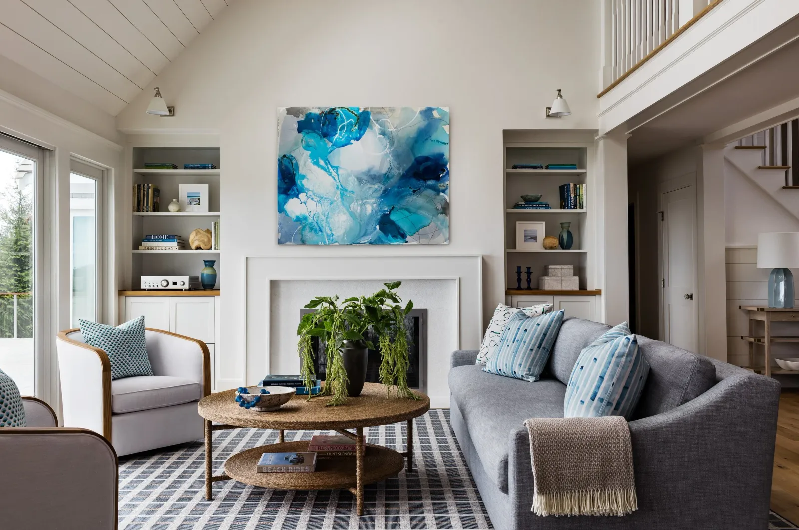 A living room with a high, vaulted ceiling, white walls, and a fireplace. The room has a gray sofa, a white armchair with a patterned pillow, a round wooden coffee table with books and a plant, and built-in shelves with books and decor. There is a la
