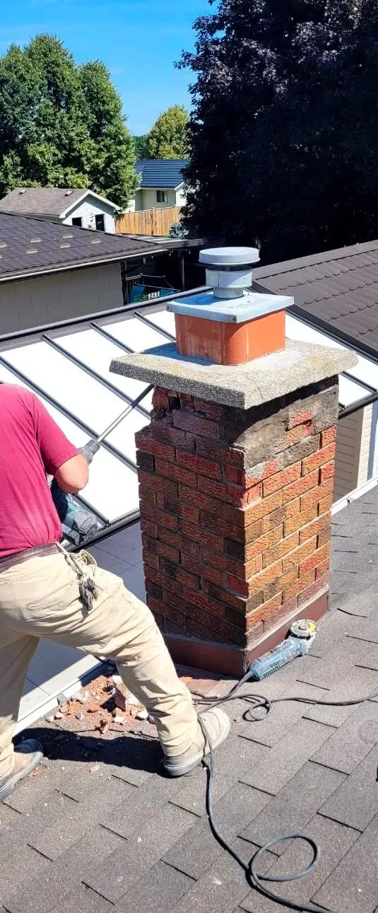 Person working on a brick chimney on a shingled rooftop, with tools and a ladder nearby.
