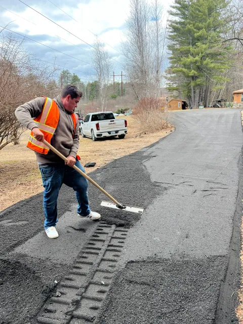 Worker in safety vest raking asphalt on a road. A truck is parked in the background, trees on either side.