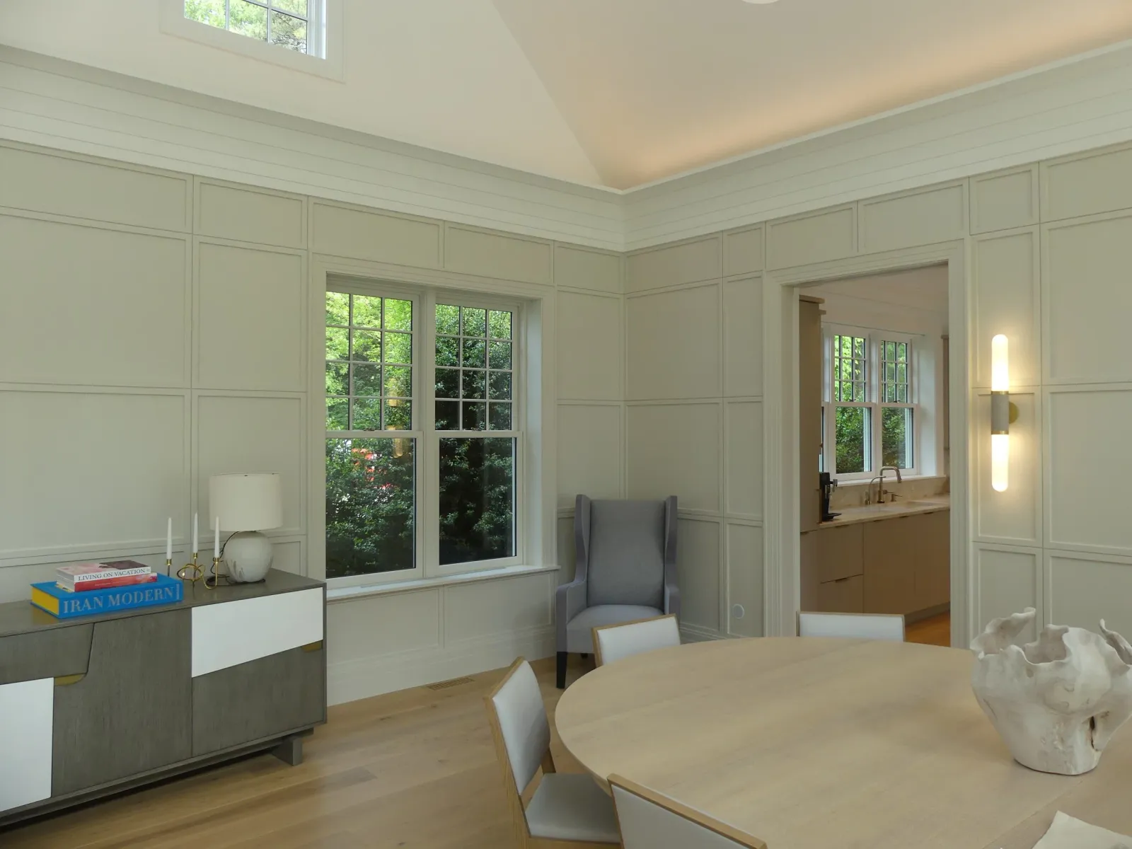A bright dining room with a round wooden table, white chairs, a sculpture in the center, a sideboard with books and a lamp, and a gray armchair near the window. There is a view of greenery outside through the large windows.