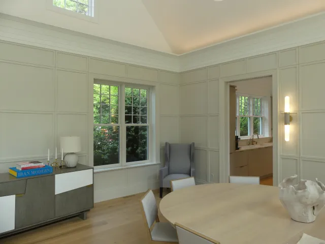 A bright dining room with a round wooden table, white chairs, a sculpture in the center, a sideboard with books and a lamp, and a gray armchair near the window. There is a view of greenery outside through the large windows.