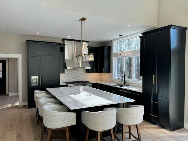 Modern kitchen with black cabinets, a white marble island, light-colored chairs, and large window with view of trees.