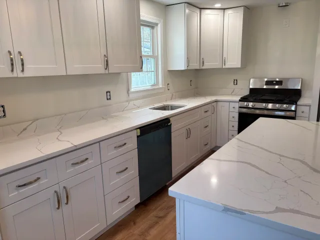 A modern kitchen with white cabinets, marble countertops, a stainless steel gas stove, and a window above the sink.