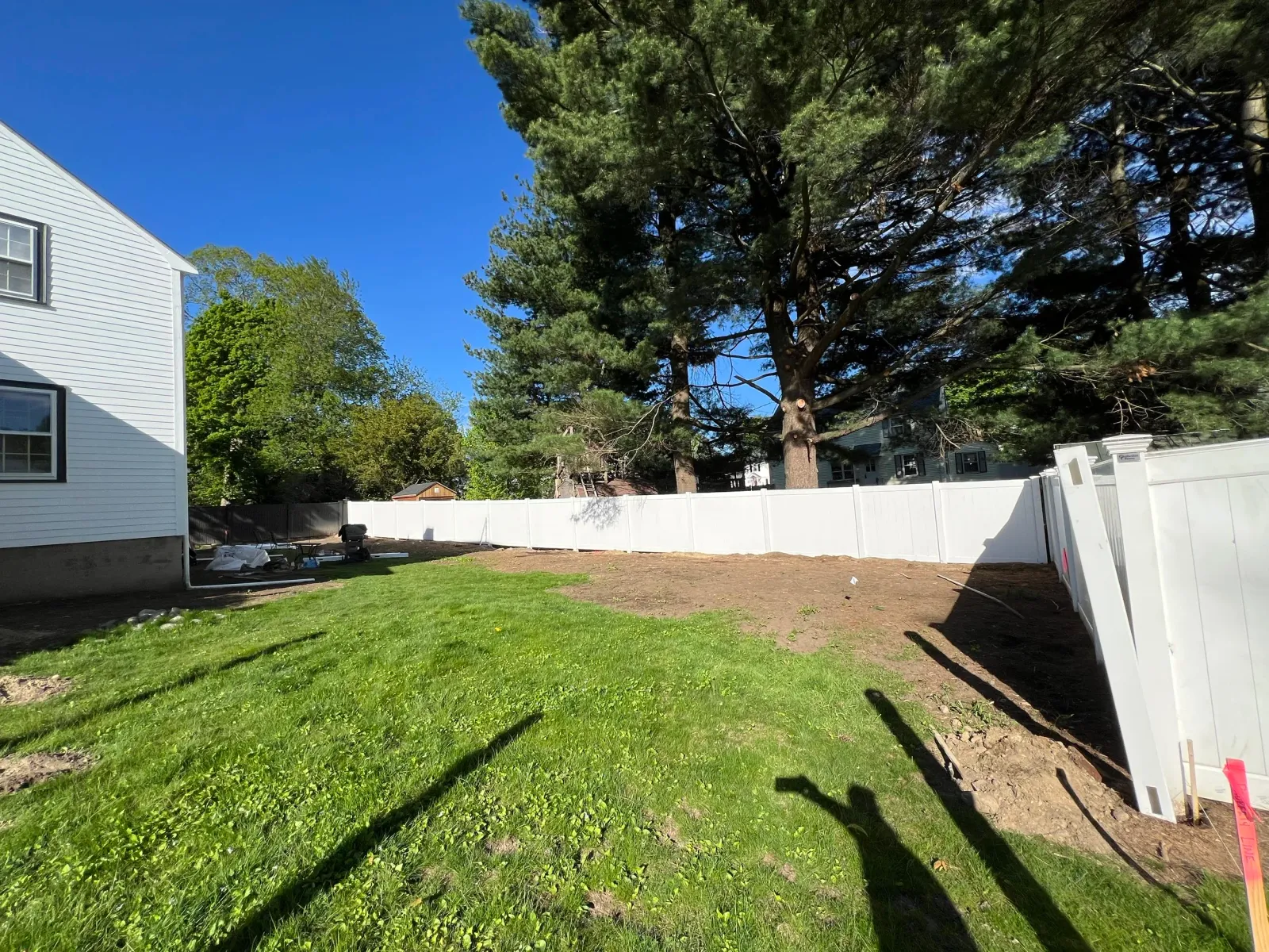 A backyard with a grassy area, a white vinyl fence, and large trees. Part of a white house is visible on the left. There are some tools and equipment on the ground near the house, and the sky is clear and blue.