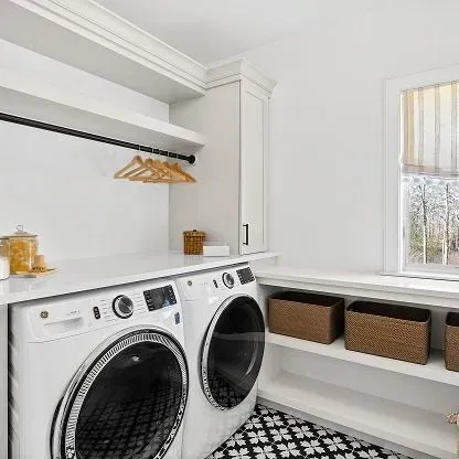 Bright laundry room with white washer and dryer, open shelves, and wicker storage baskets near a window.
