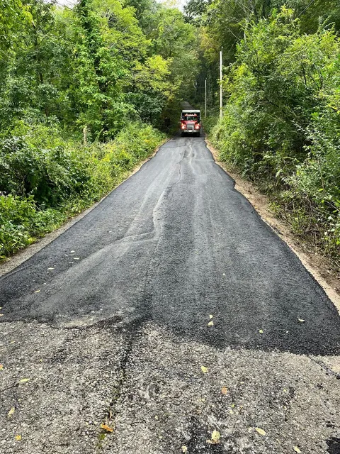 Newly paved road leading into a tree-lined area; a bus is visible in the distance.