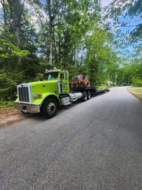Lime green semi-truck with flatbed trailer on asphalt road, carrying construction equipment, trees in background.