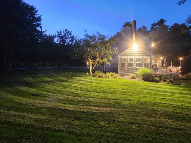 A well-maintained backyard with freshly mowed green grass, a small tree, and shrubs near a house. The house has a porch with lights, and the scene is illuminated by outdoor porch lights at dusk or night.