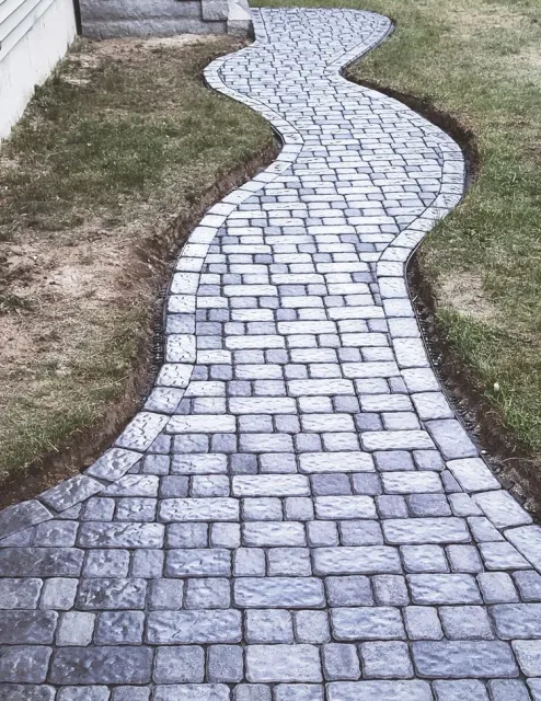 Curving stone pathway in a yard, bordered by grass and a building wall.