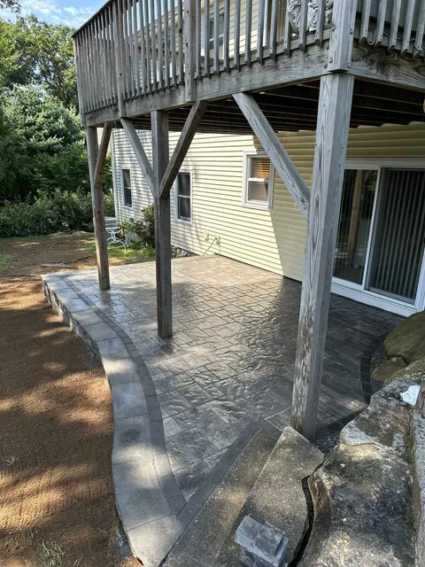 A gray stone patio sits beneath a wooden elevated deck attached to a light yellow house.