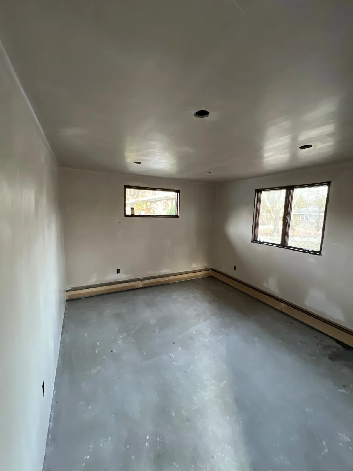 Empty room under construction with unfinished concrete floor, white walls, two small windows, and ceiling with multiple light fixture holes.