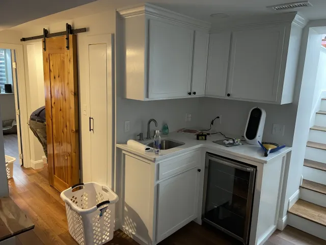 A small kitchen area with white cabinets, a mini wine refrigerator, and a countertop with tools and cleaning supplies. There is a laundry basket on the wooden floor and a sliding barn door leading to another room. A staircase is visible to the right.