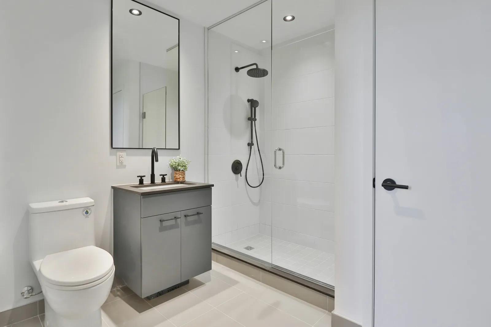 Modern bathroom with a toilet, gray vanity with black faucet, potted plant, large mirror, and glass-enclosed shower with black fixtures.