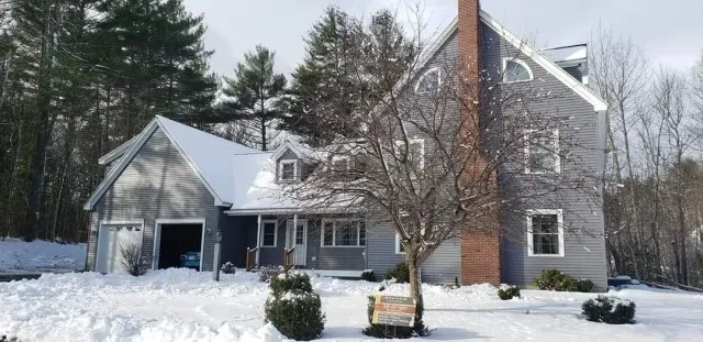 A large house with gray siding and a brick chimney in a snowy landscape, with a tree and bushes in front.