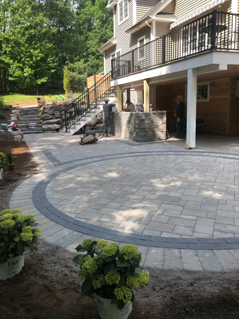A newly paved outdoor patio with a circular pattern, surrounded by landscaping, and a construction crew working on a stone wall beneath a house with a deck.