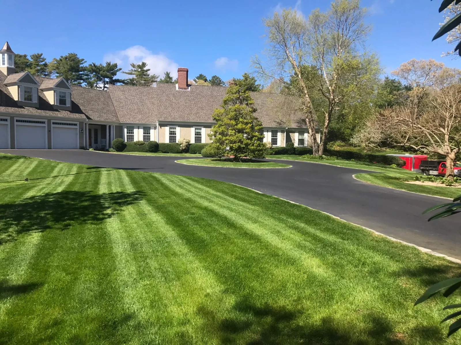 Large suburban house with a well-maintained green lawn, asphalt driveway, multiple garage doors, surrounding trees, and a blue sky.