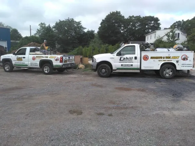Two white utility trucks parked on gravel near trees and a house