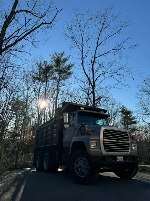 A large gray commercial truck parked on the side of a paved road surrounded by leafless trees with the sun shining through.