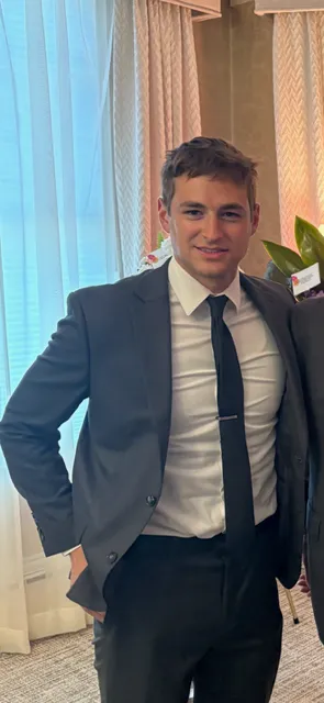 Young man in a navy suit and tie standing indoors near a window with curtains.