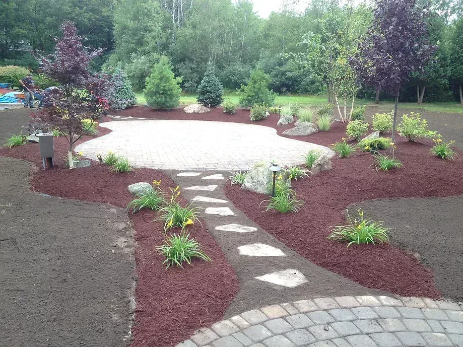 A stone path leads to a circular patio surrounded by dark red mulch and various shrubs in a residential yard.