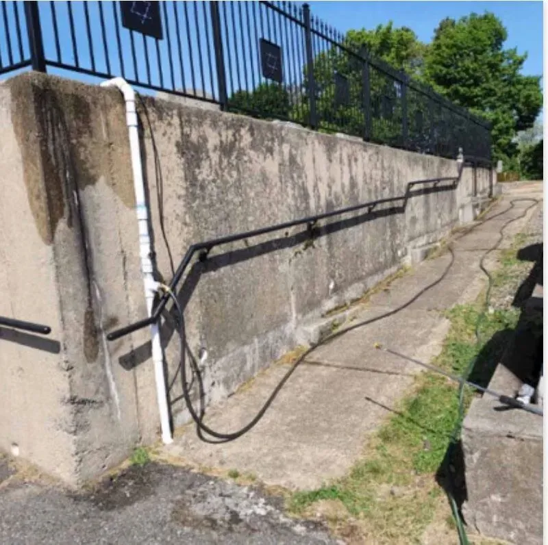 Concrete wall with black railing, black fence in the background, white pipes, and a walkway along the wall.